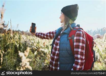 Young woman taking selfie with smartphone during vacation trip in mountains. Woman with backpack hiking through tall grass along path on meadow. Spending summer vacation close to nature