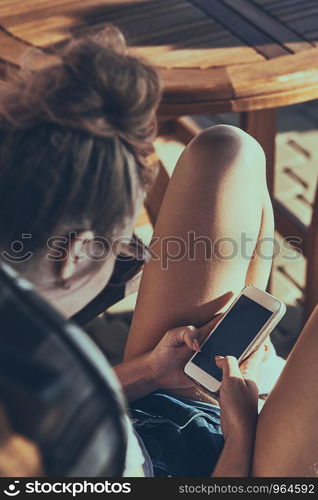 Young woman talking on a mobile phone sitting in a chair outdoors on patio wearing a sunglasses. Real people, authentic situations