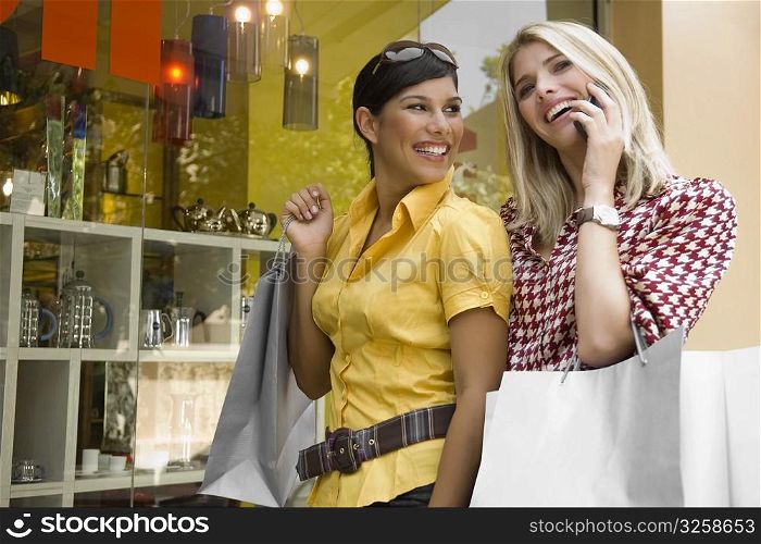 Young woman talking on a mobile phone with another young woman holding a shopping bag beside her
