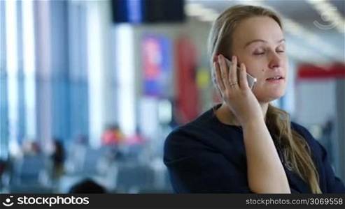 Young woman talking on the phone in the waiting room of airport or station, she giving information about her travel. Defocused interior in background
