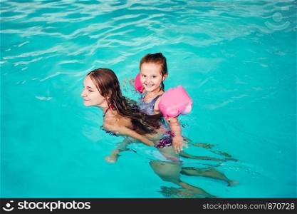 Young woman teaching to swim her younger sister and playing with her in swimming pool. Candid people, real moments, authentic situations