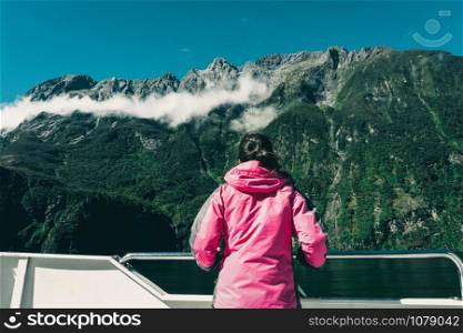 Young woman tourist looks at fjord scenery from the ship deck in Milford Sound in Fiordland National Park, South Island of New Zealand. Boat cruise in Milford Sound is main activity for most tourist.