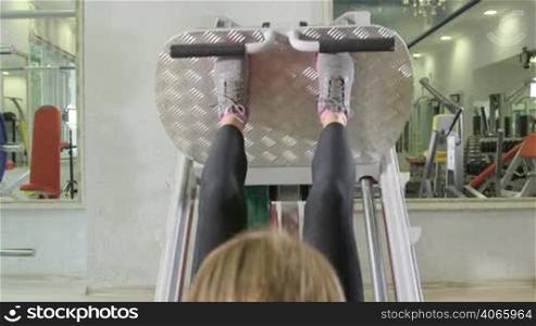 Young woman training in health fitness club on leg press machine, closeup rear view