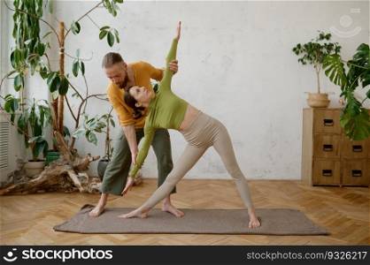 Young woman training yoga with male instructor indoors. Man teacher helping female student to make Trikonsana asana. Young woman training yoga with male instructor indoors