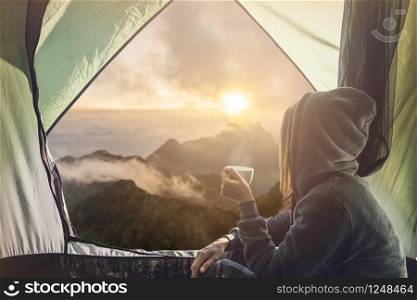 Young woman traveler sitting in the tent, relaxing and drinking coffee with beautiful sunrise