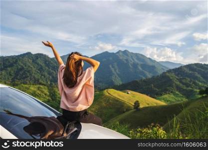 Young woman traveler sitting on a car watching a beautiful mountain view while travel driving road trip on vacation