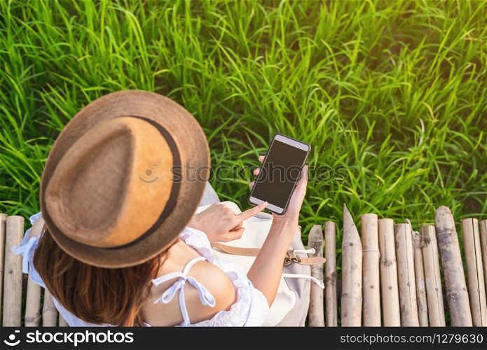Young woman traveler sitting on bamboo path and using smart phone at green paddy field