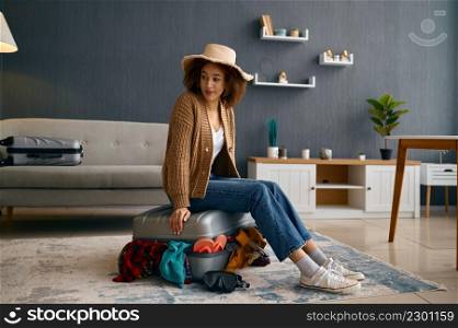Young woman traveler sitting on suitcase lid with clothes excess and dreaming about future vacation. Woman sitting on suitcase with clothes excess