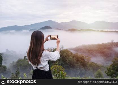 Young woman traveler taking photo with smart phone at sea of mist and sunrise over the mountain