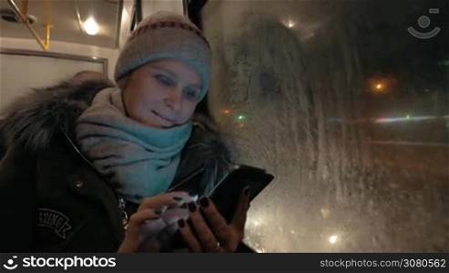 Young woman traveling in the city by bus in winter evening. She browsing web on smart phone and smiling when looking at screen