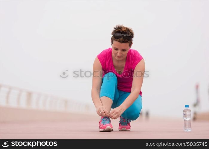 Young woman tying shoelaces on sneakers on a promenade. Standing next to a bottle of water. Exercise outdoors