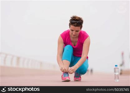 Young woman tying shoelaces on sneakers on a promenade. Standing next to a bottle of water. Exercise outdoors