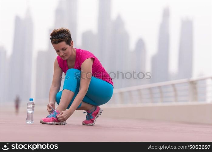 Young woman tying shoelaces on sneakers on a promenade with a big city in the background. Standing next to a bottle of water. Exercise outdoors