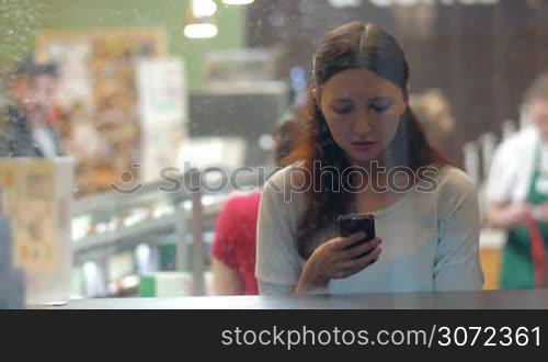 Young woman typing sms or chatting in social network service on mobile phone in a cafe. View through the glass in rainy evening