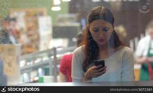 Young woman typing sms or chatting in social network service on mobile phone in a cafe. View through the glass in rainy evening