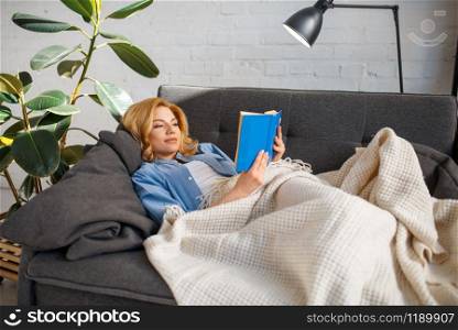 Young woman under a blanket reading a book on cozy yellow couch, living room in white tones on background. Attractive female person with magazine sitting on sofa. Young woman under a blanket reading book on couch