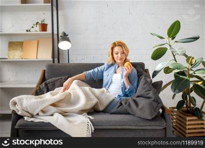 Young woman under a blanket reading a book on cozy yellow couch, living room in white tones on background. Young woman under a blanket reading book on couch