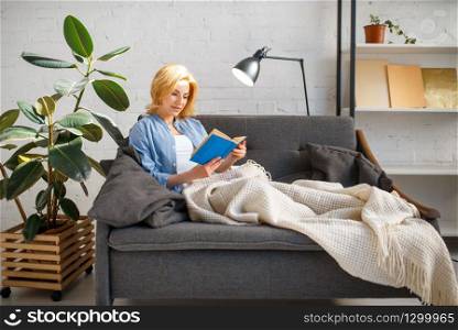 Young woman under a blanket reading a book on cozy yellow couch, living room in white tones on background. Attractive female person with magazine sitting on sofa at home