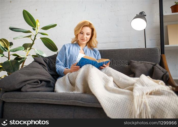 Young woman under a blanket reading a book on cozy yellow couch, living room in white tones on background. Attractive female person with magazine sitting on sofa at home. Young woman under a blanket reading book on couch