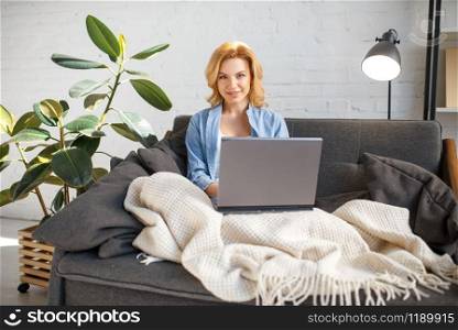 Young woman under a blanket using laptop on cozy yellow couch, living room in white tones on background. Attractive female person with magazine sitting on sofa at home. Young woman under a blanket using laptop on couch