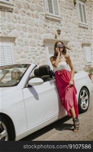 Young woman uses a mobile phone and stands next to cabriolet in the summer day