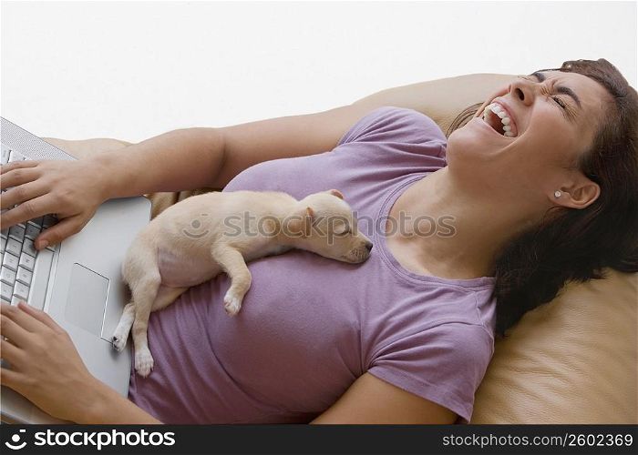 Young woman using a laptop and laughing with a puppy lying on her