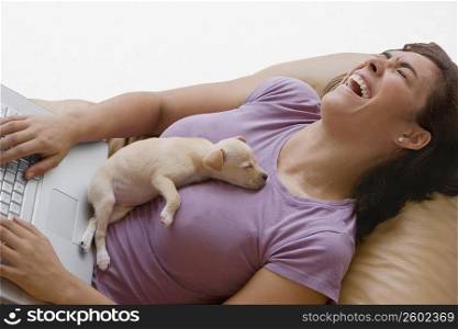 Young woman using a laptop and laughing with a puppy lying on her