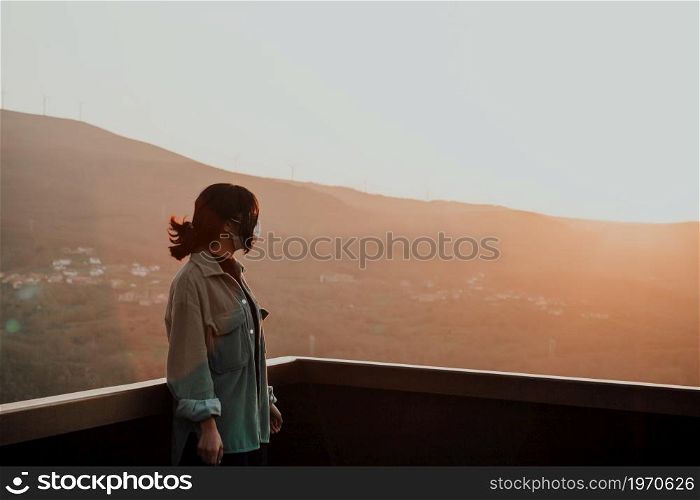 Young woman using a mask looking the mountains from a balcony during a super colorful sunset with copy space, freedom and wellness concept