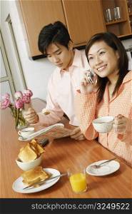Young woman using a mobile phone and a young man reading a newspaper at a kitchen counter