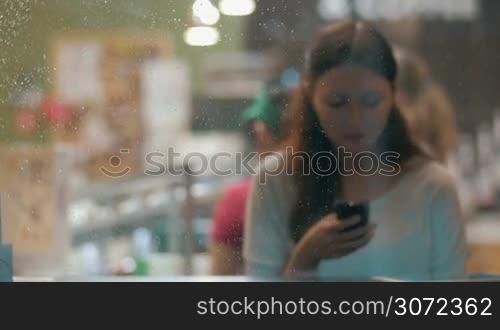 Young woman using cell phone to type text message or communicate in social media. She sitting alone in the cafe in the evening, view through the wet glass