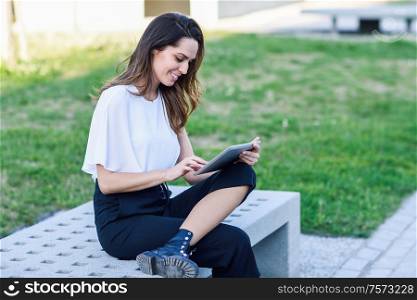 Young woman using digital tablet sitting outdoors in urban background.. Middle-age woman using digital tablet sitting outdoors in urban background.