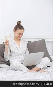 Young woman using her laptop while relaxing in bed and drinking wine. To stay home. Online study. Remote work. Young woman using her laptop while relaxing in bed and drinking wine. To stay home. Online study. Remote work.