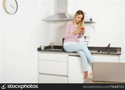 Young woman using her smartphone sitting in the kitchen at home. Thinking beautiful woman using her smartphone sitting in the kitchen at home