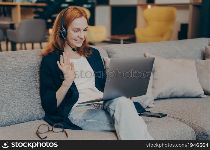 Young woman using laptop and wireless headset for internet video call with friends, waving hand in greeting gesture, broadly smiling while looking on computer screen. Online communications concept. Young woman using laptop and headset for internet video call waving hand in greeting gesture