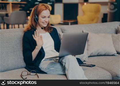Young woman using laptop and wireless headset for internet video call with friends, waving hand in greeting gesture, broadly smiling while looking on computer screen. Online communications concept. Young woman using laptop and headset for internet video call waving hand in greeting gesture