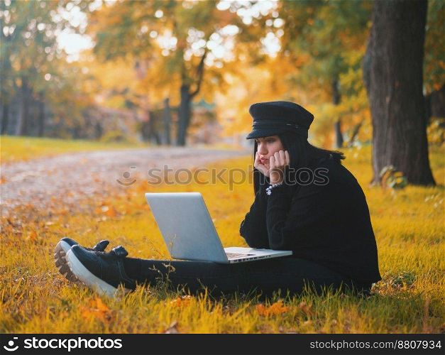 Young woman using laptop outdoor sitting on yellow grass and smiling. Girl using digital tablet pc in the park. Student using tablet after school.. Sad young woman using laptop outdoor sitting on yellow grass. Girl using digital tablet pc in the autumn park. Trendy student in fashion hat using tablet after school.