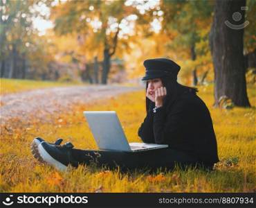 Young woman using laptop outdoor sitting on yellow grass and smiling. Girl using digital tablet pc in the park. Student using tablet after school.. Sad young woman using laptop outdoor sitting on yellow grass. Girl using digital tablet pc in the autumn park. Trendy student in fashion hat using tablet after school.