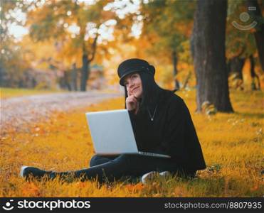 Young woman using laptop outdoor sitting on yellow grass and smiling. Girl using digital tablet pc in the park. Student using tablet after school.. Pensive young trendy woman using laptop outdoor sitting on yellow grass and smiling. Girl using digital tablet pc in the park. Student in fashion cap using tablet after school.