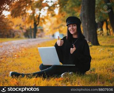 Young woman using laptop outdoor sitting on yellow grass and smiling. Girl using digital tablet pc in the park. Student using tablet after school.. Young woman in cap using laptop outdoor sitting on yellow grass and smiling. Girl using digital tablet pc in the park. Student using tablet after school and showing ok sign.