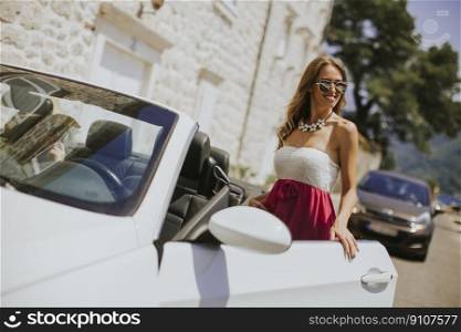 Young woman using mobile phone and standing by cabriolet car at hot summer day