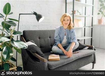 Young woman using mobile phone on cozy black couch, living room in white tones and pot with flower on background. Attractive female person sitting on sofa at home. Young woman using mobile phone on cozy black couch