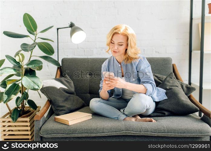 Young woman using mobile phone on cozy black couch, living room in white tones and pot with flower on background. Attractive female person sitting on sofa at home