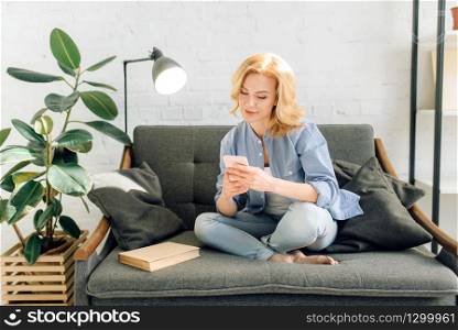 Young woman using mobile phone on cozy black couch, living room in white tones and pot with flower on background. Attractive female person sitting on sofa at home