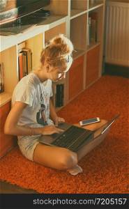 Young woman using portable computer and mobile phone, sitting on a floor, learning online at home. Candid people, real moments, authentic situations