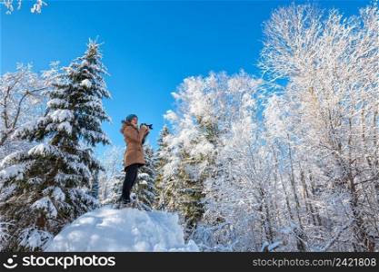 Young woman walking in winter forest and taking photos among snow-covered trees