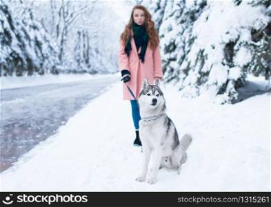 Young woman walks in the park with siberian husky, snowy forest on background. Cute girl with charming dog