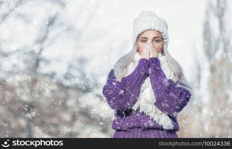 Young woman warming her hands, wearing warm clothing on snowing winter day outdoors. Cute girl feeling cold. Winter concept. Copy space for text.