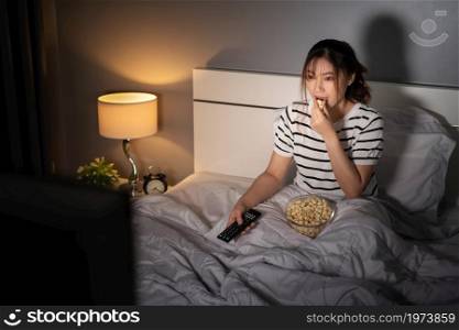 young woman watching TV and eating popcorn on a bed at night