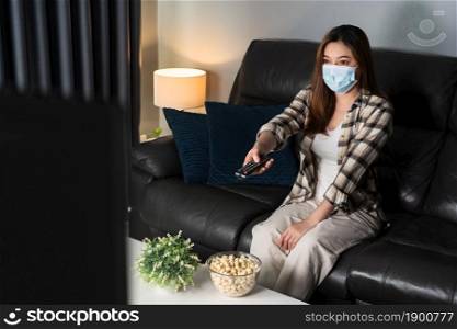 young woman watching TV on sofa and wearing medical mask to protect coronavirus (Covid-19)