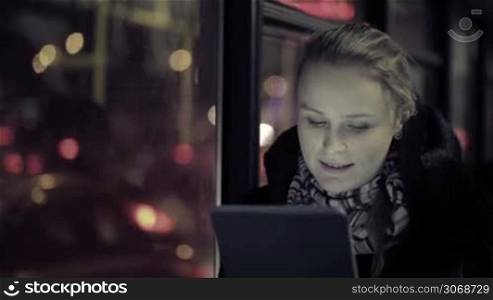 Young woman watching video on touchpad during evening ride in the bus. Defocused city traffic in the bus window